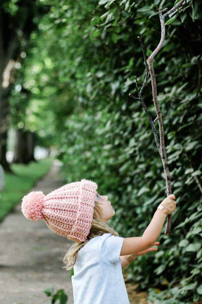 Young girl in a pink crochet hat holding a tree branch.