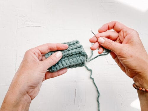 A woman's hands holding a tapestry needle and a crochet rectangle folded into a tube shape.