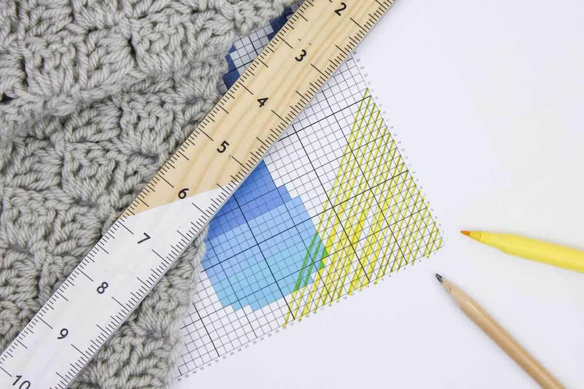 A close-up shot of a C2C pattern guide with a marker, pencil, and ruler with a gray crochet blanket on a white background.