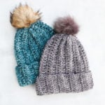 Two chunky crochet beanies with fur pom poms laying on top of a white background.